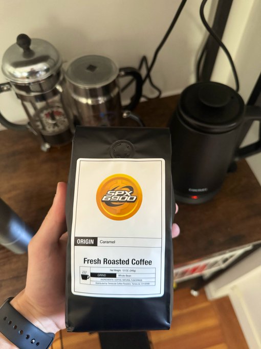 The image features a person holding up a bag of coffee beans, which has the text "Fresh Roasted Coffee" printed on it. The background includes a coffee maker and a blender, suggesting that this scene takes place in a kitchen or a similar setting where coffee is being prepared. The caption at the bottom of the photo reads "Aeons support aeons Paid for with #SPX6900 obviously". This implies that the person holding up the bag of coffee beans might be promoting a product or service related to coffee, possibly as part of an advertisement campaign.