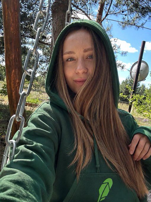 A young woman with long hair wearing a green hoodie takes a selfie while sitting on a swing with metal chains, with trees and a partly cloudy sky in the background.