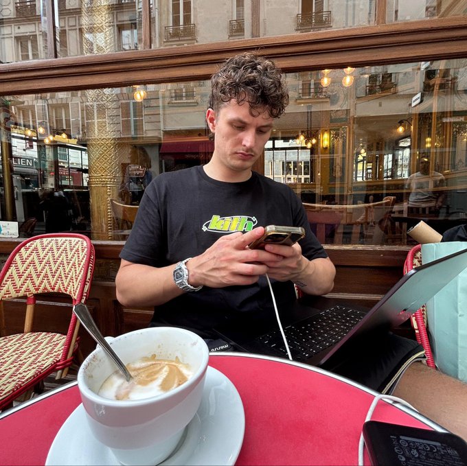 A young man with curly hair sits at an outdoor café table looking at his phone, with a cappuccino, laptop, and a classical building visible through the window behind him.
