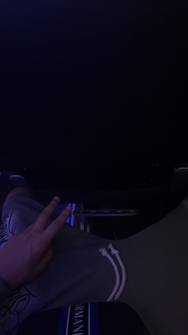 A dimly lit photo showing a person's hand making a peace sign, wearing a gray hoodie. In the background, there appears to be the dark silhouette of an object, possibly an arcade game or machine.