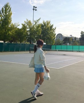 A young woman in a light jacket and blue skirt walking on a tennis court carrying a racket and shopping bags.