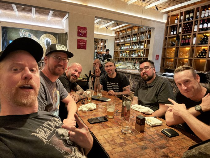 A group selfie of seven men gathered around a table at a wine bar or restaurant, smiling and posing with drinks amid a background of extensive wine racks and bottles.