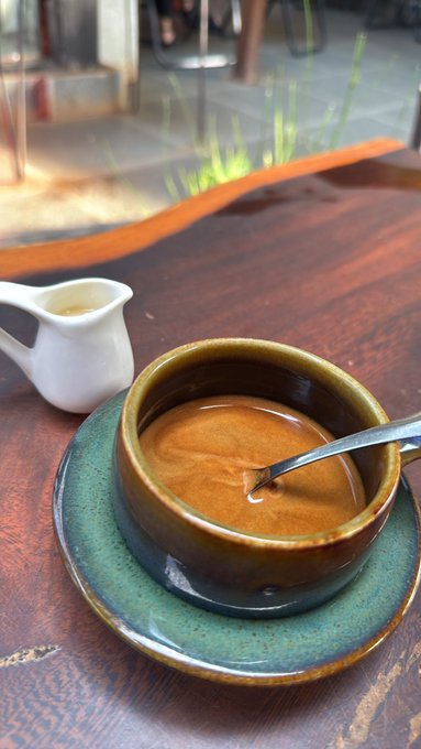 A close-up photo of a small green ceramic cup filled with espresso on a matching saucer, sitting on a wooden table beside a small white creamer pitcher.