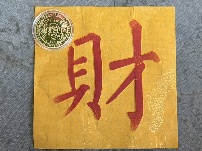 A yellow paper envelope with red Chinese characters '財' (wealth) printed on it sits next to a coin labeled 'TST', with the token details '5.7', '195', and 'BLOCK' visible on its face.