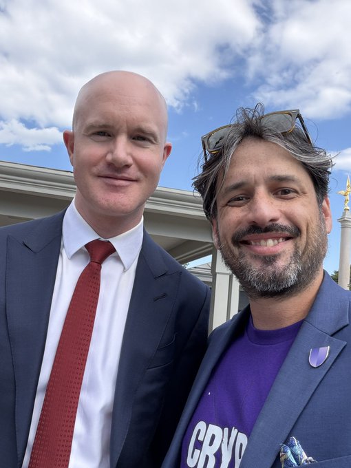 A selfie of two smiling men outdoors against a partly cloudy sky; the man on the right is wearing a purple t-shirt with the partial word 'CRYP' visible, under a blazer with a purple pin.