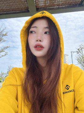 A selfie of a young woman wearing a yellow Binance hoodie with the brand logo visible on the chest and sleeve, taken outdoors under a canopy.