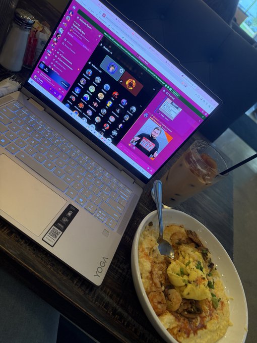 A Lenovo Yoga laptop sits on a wooden table displaying what appears to be a Discord call interface, next to a plate of shrimp and rice with an iced coffee drink.