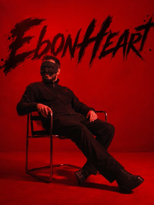 Promotional photo of a man in all black clothing wearing a black mask, seated in a chair against a red background with "EBON HEART" written in large black brushstroke text above him.