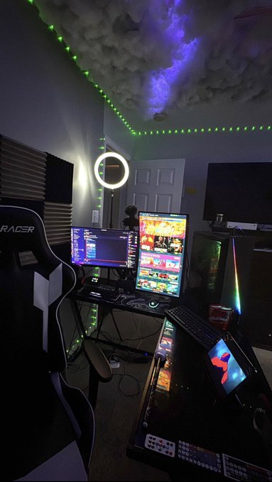 A gaming setup features a racing chair labeled "RACER," dual monitors, a ring light, green LED strip lighting, and a custom monitor displaying colorful content under cloud-like ceiling decor.