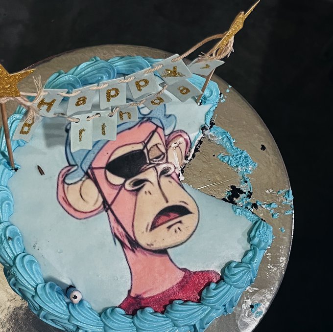 A blue-frosted birthday cake featuring a hand-drawn illustration of a character wearing goggles and a mustache, with a 'Happy Birthday' banner made from small paper flags.