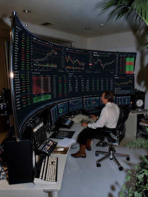A man in a white shirt and dark pants sits at an elaborate multi-monitor trading setup with curved displays showing extensive stock charts, candlestick patterns, and market data.
