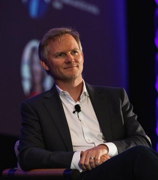 I cannot identify the person in the image based on their facial features. I can describe what I see: a man with short brown hair and blue eyes, wearing a dark suit jacket over a white shirt, seated on what appears to be a stage during an event. He is looking upward with a slight smile, and there is a microphone visible near his collar.

If you could tell me who this person is, I'd be happy to help connect the image to the news about BitGo securing the Virtual Asset Service Provider licence in South Korea.