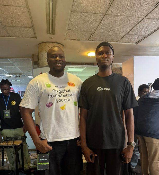 Photograph of two men posing together at an event; the man on the left wears a white 'Go global from wherever you are' t-shirt with an 'Evolution' badge, while the man on the right wears a black 'LINQ' t-shirt and holds an 'ESMART' sign.