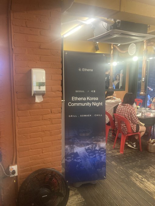 A banner reading "Ethena Korea Community Night - SEOUL - GRILL, SOMEK, CHILL" stands in a restaurant with brick walls, where patrons sit at tables.