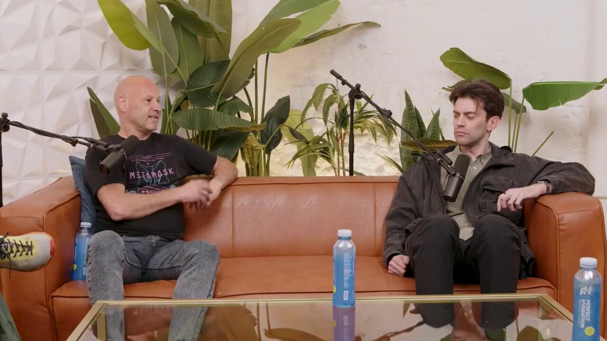 The image shows two men sitting on a couch, engaged in conversation. One man has his arm around the other's shoulder as they talk. There are several water bottles scattered around them, indicating that they might be enjoying some refreshments during their discussion. The background features a wall with plants and a coffee table, creating a relaxed atmosphere for their conversation.