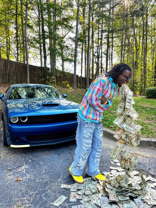 A person in a colorful plaid shirt, ripped jeans, and yellow shoes stands next to a blue Dodge Challenger, appearing to spit out a large stream of US dollar bills.