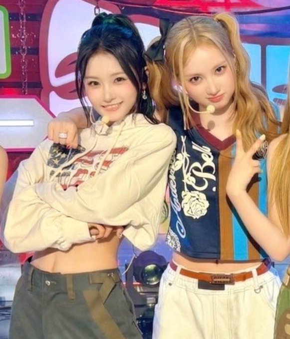 Two young women posing together on a colorful stage, one with dark hair in a cream crop top and the other with blonde pigtails in a blue vest with rose embroidery.