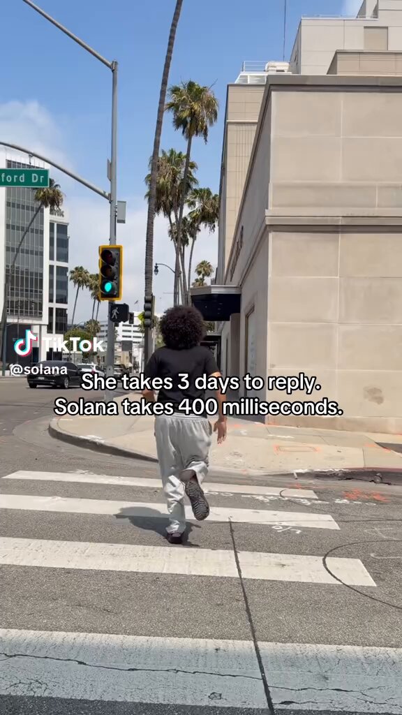 The image shows a woman walking across a street, wearing white pants and a black shirt. She appears to be looking at her phone while crossing the road. The photo has been shared on social media with the caption "I'm just saying go follow Solana on TikTok man tiktok.com/ZP8sMFEKc/". This means that the woman in the image is encouraging others to follow a particular person or account, likely Solana, on TikTok.
