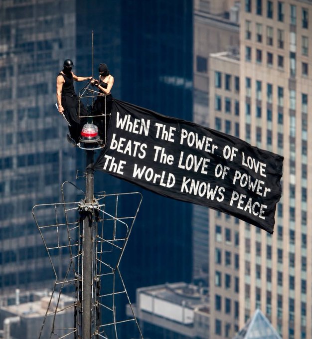 Two masked activists perch atop a tall antenna structure against a city skyline, unfurling a large black banner that reads "When the power of love beats the love of power the world knows peace," likely capturing the rooftop proposal moment of daredevil artist Angela Nikolau and her partner.