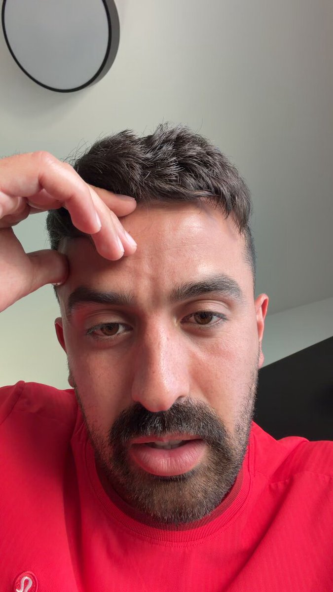 The image features a man with a beard wearing a red shirt, who has his hand on his head as if he's scratching an itch or feeling unwell. The background of the photo shows a white wall, which contrasts with the man's red shirt. There is also a small circular light fixture in the top left corner of the image.
