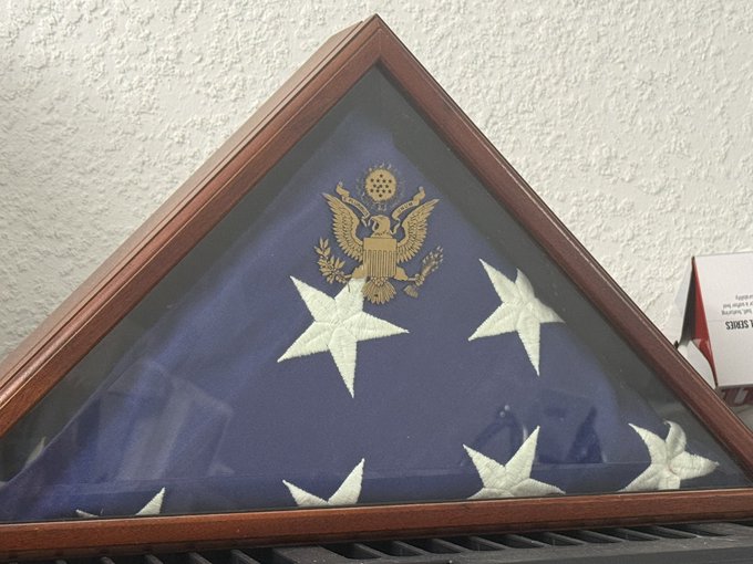 A wooden triangular display case holds a folded American flag beneath the gold Great Seal of the United States eagle emblem.