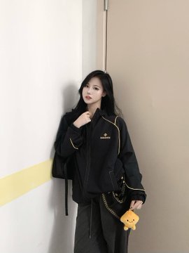 Photo of a woman with long dark hair wearing a black jacket and holding a small bear-shaped bag, standing against a white wall.