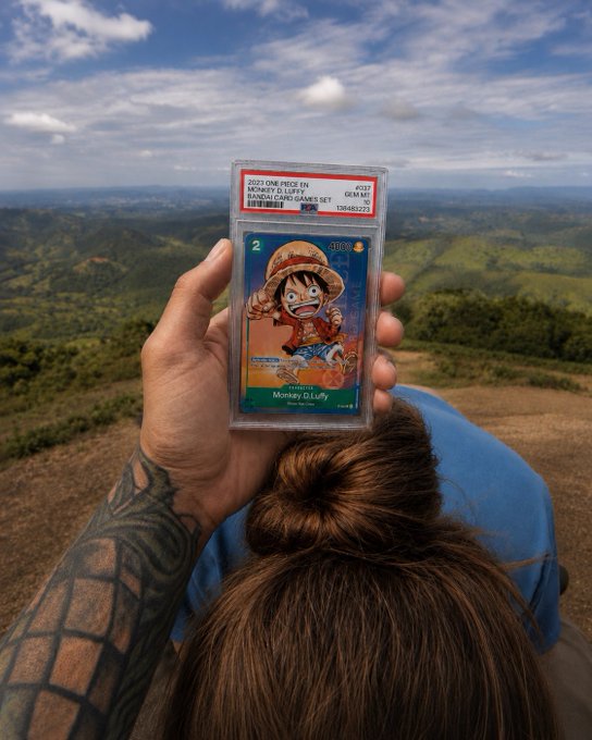A hand holds up a PSA-graded 2023 One Piece card featuring Monkey D. Luffy (#337, Gem Mint 10) against a scenic mountain backdrop, illustrating the post's choice between a high-value collectible card and 0.5 ETH in cryptocurrency.