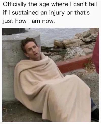 The image is a screenshot of a social media post with the caption: "Officially the age where I can't tell if I sustained an injury or that's just how I am now." Below the text is a photo of a man wrapped in a blanket, sitting against a stone wall with his head slumped over, looking exhausted or defeated.