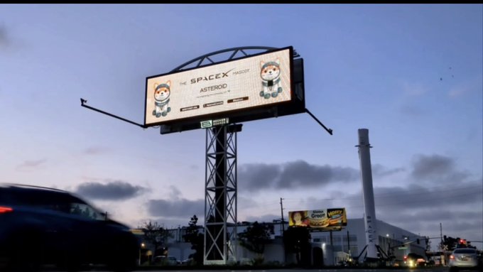 A billboard at dusk promotes "THE SPACEX MASCOT ASTEROID" featuring astronaut characters, positioned near the SpaceX headquarters. In the background, the white SpaceX smokestack is visible against a cloudy sky, tying the advertisement directly to the company's location.