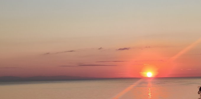 Serene photograph of a sunset over a calm ocean, with the orange sun low on the horizon casting reflections on the water, distant mountains visible to the left, and a few wispy clouds in the pink and orange sky.