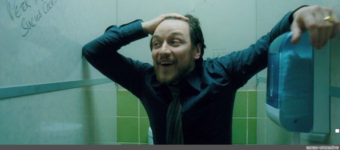 A man with a beard and dark shirt smiles broadly with his arms raised in a green-tiled bathroom, appearing to be from a film still.