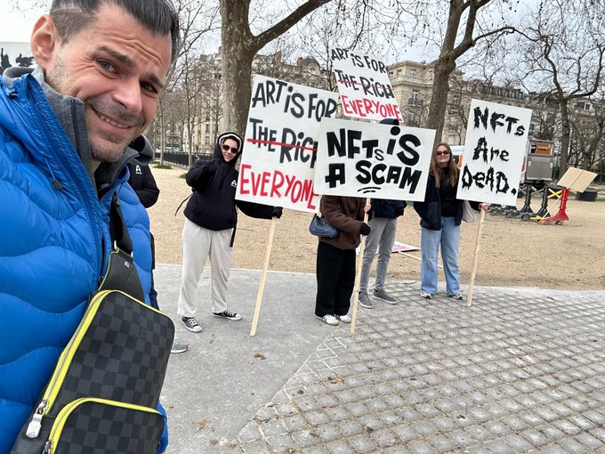 A man takes a selfie with protesters holding signs reading 'ART IS FOR THE RIGHT EVERYONE' and 'NFTs IS A SCAM', 'NFTs Are Dead.'