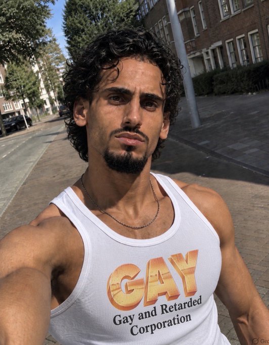 A man in a white tank top takes a selfie on a street, with his shirt reading "GAY - Gay and Retarded Corporation," playfully referencing the ticker mentioned in the post about the $GAY cryptocurrency's minor 0.15% gain.