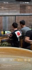 A blurry, vertical video clip of two people standing outdoors near a wooden structure, possibly interacting or struggling, with video playback controls visible at the bottom.