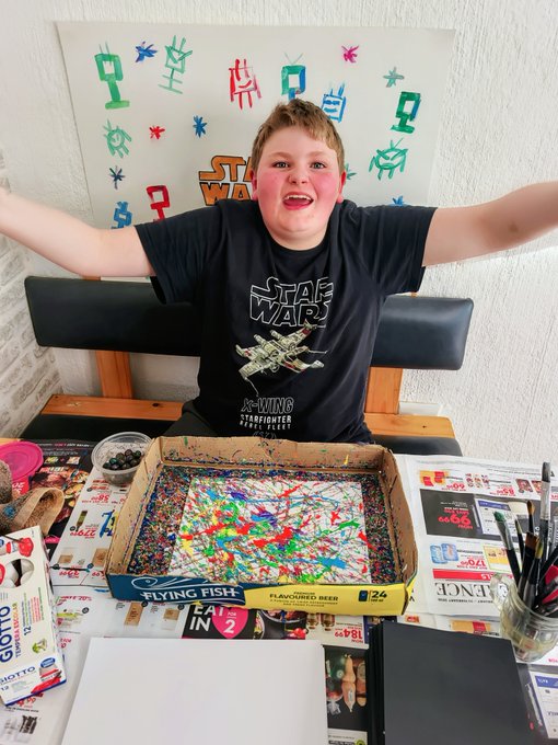 A beaming boy in a Star Wars X-Wing Starfighter t-shirt sits proudly behind a paint-covered pizza box canvas splattered with vibrant colors, surrounded by Star Wars-themed artwork and art supplies. The colorful abstract piece perfectly captures the "rainbow" NFT theme mentioned in the post, with his enthusiastic pose celebrating the creative crypto-inspired art.