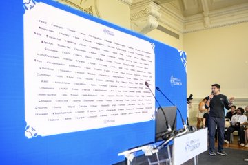 A conference or event setting where a man in a black shirt is presenting in front of a large blue backdrop covered with numerous sponsor logos.