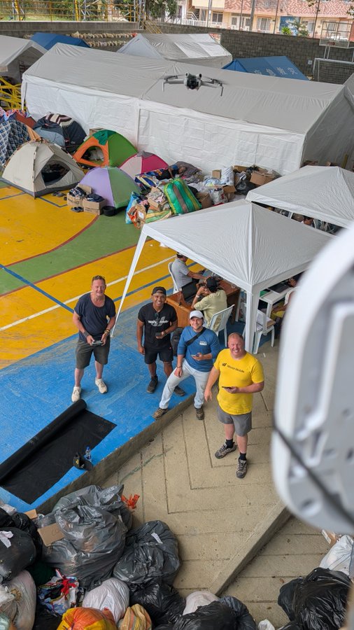 An aerial view of people gathered under white tents and umbrellas on a colorful outdoor court, with piles of trash bags nearby, possibly indicating a relief or encampment site.