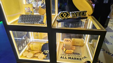 A WEEX branded display case showcases merchandise including a yellow "ONE ACCOUNT, ALL MARKETS" box, exclusive TOKEN2049 collectibles, and a WEEX logo plaque. The image ties to the post by highlighting WEEX's presence at TOKEN2049 Day 2, where they featured exclusive merch from Pudgy Penguins alongside an AI trading demo.