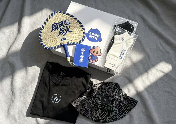The image features a white box with various items, including a black shirt, hat, and fan. The box also contains a small stuffed animal and a card. The objects are arranged in such a way that they appear to be placed on top of each other or stacked together. The text "Love HNX" is visible in the image, which could potentially be related to an online store selling these items.
