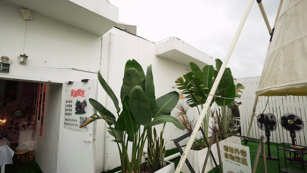 The image shows a white building with a large green plant in front of it. The building has a sign that reads "1/ BBQ, Debate & Future Narratives: Team1 AI & RWA Night Successfully Concluded!". In the background, there is an open area where people are gathered for a BBQ event. A person can be seen standing near the building with the green plant in front of it. The scene also includes some chairs and a dining table placed outside on the grassy area.