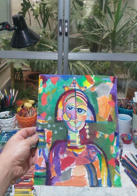 A hand holds up a vibrant, abstract mixed-media portrait painting featuring bold colors, with art supplies like colored pencils and paints visible on the desk and green plants in the background.
