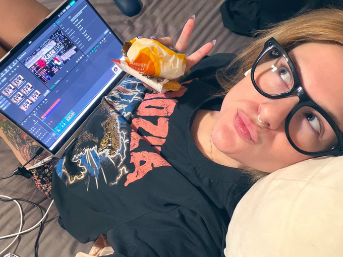 A young woman with glasses and a nose ring lying down, holding an open bag of Cheetos while working on video editing software (showing a timeline) on a laptop.