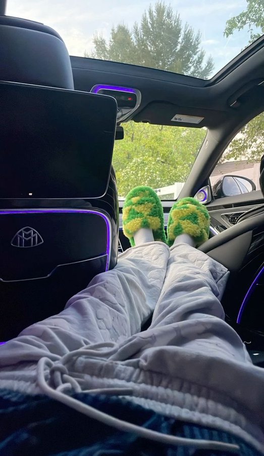 A view from inside a car with a sunroof, showing legs stretched out wearing white pants and fuzzy green and yellow striped slippers, with trees visible through the windows.