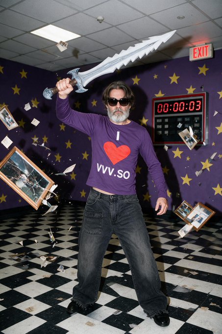 A man wearing sunglasses and a purple shirt reading 'I [heart] vvv.so' holds a large silver sword aloft in a room with star-patterned walls and a checkered floor, with shattered picture frames scattered around.