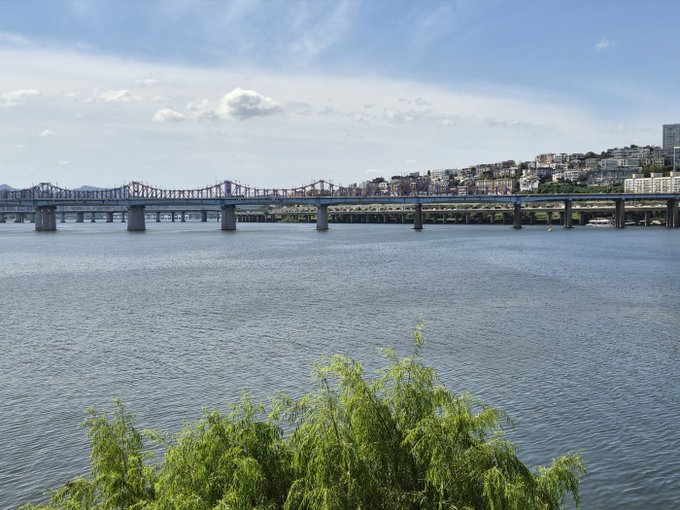 A scenic daytime view shows a multi-tiered bridge spanning a wide river, with a city skyline in the distance and green trees in the foreground.