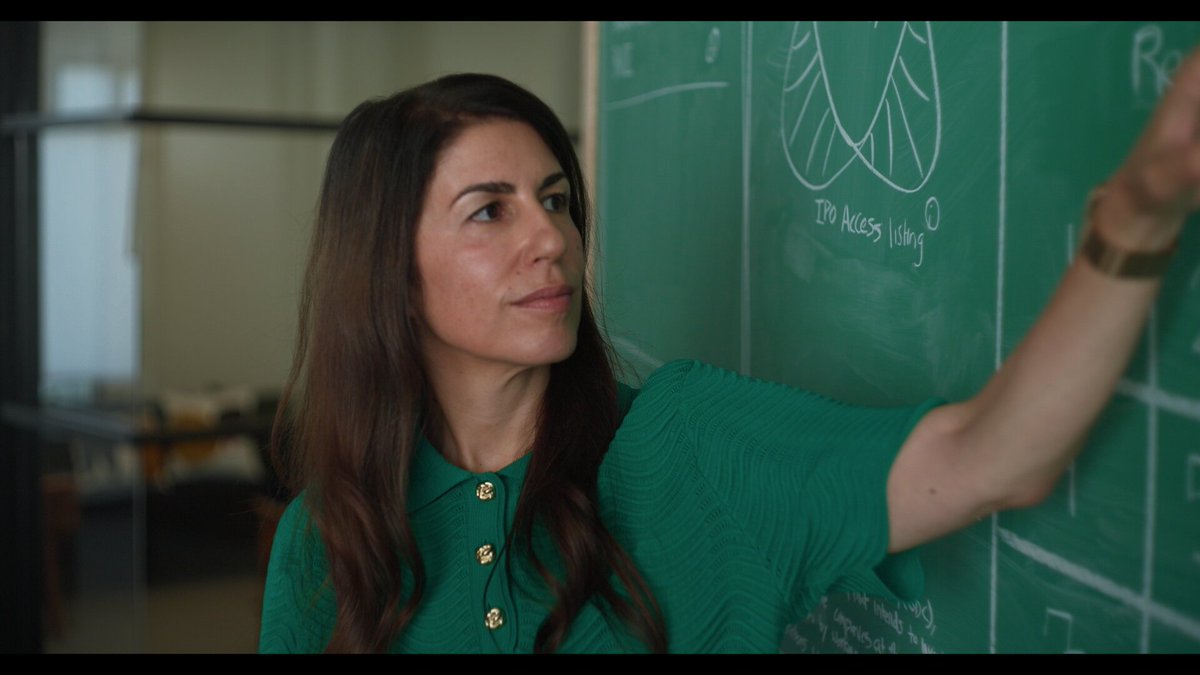 A woman with long brown hair wearing a green buttoned shirt stands in a classroom, writing on a green chalkboard.