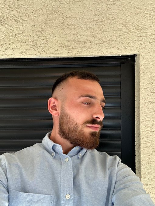 A man with a short buzz cut and full beard, wearing a light blue button-down shirt, poses in front of a black roll-up door and textured cream wall. The casual selfie matches the cheerful "Gm friends" greeting with a confident, morning-ready vibe.