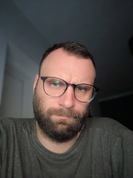 A close-up selfie of a bearded man wearing glasses and a dark speckled t-shirt, looking directly at the camera in a dimly lit indoor setting.