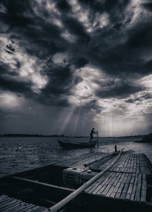 A solitary figure stands in a small boat beneath dramatic sunbeams piercing through dark clouds over calm waters, with a wooden raft in the foreground—visually embodying the post's "silver lining" metaphor. Shot on a Nikon D7200 with a Tokina 11-16mm lens, the black and white composition contrasts the moody, stormy sky with rays of hope breaking through.