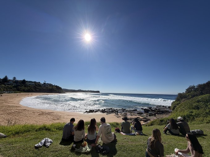 A group of people sits on the grass overlooking a scenic beach cove with bright sun and blue sky, watching the waves.